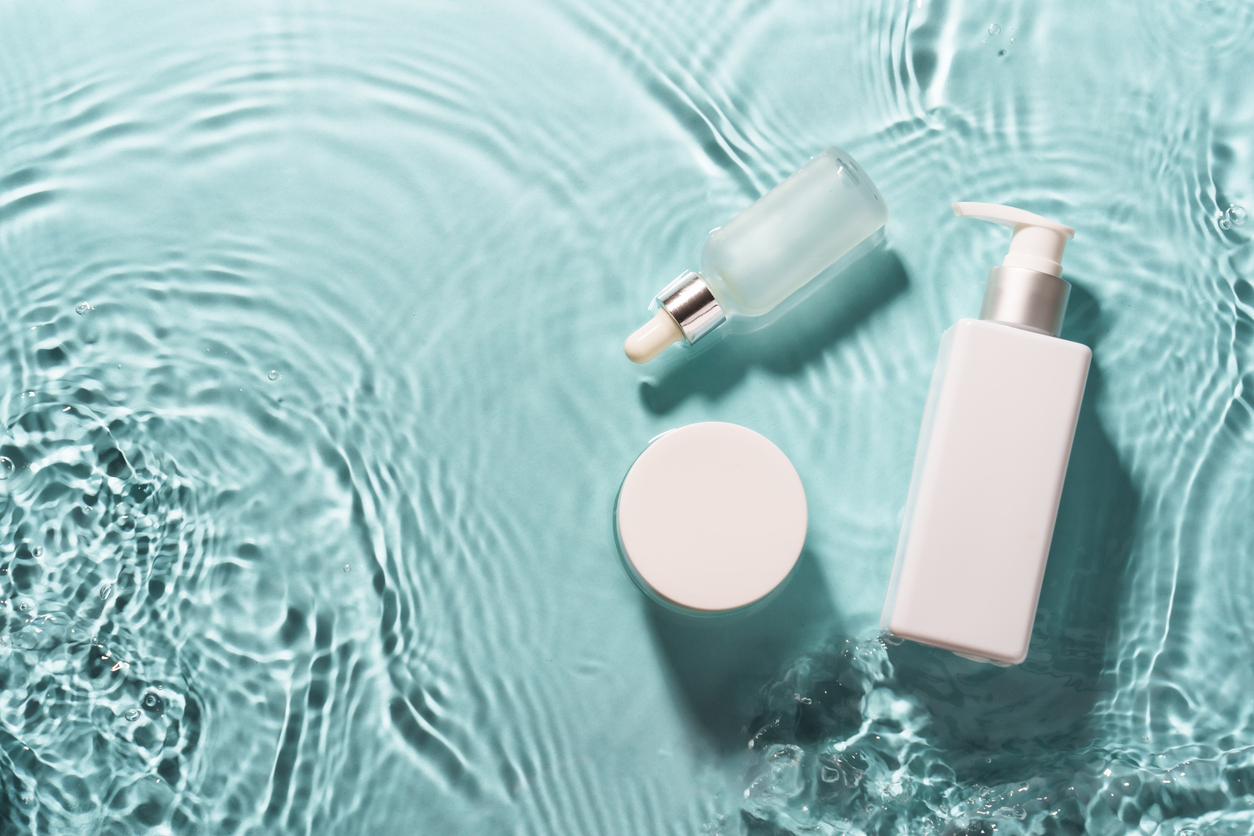 Skincare dropper bottle, jar and pump bottle resting on rippled turquoise water
